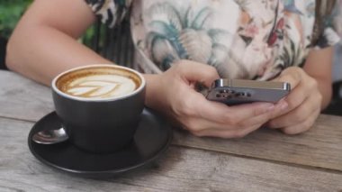 Pembe tırnaklı kız haberleri yeni moda bir telefonla izliyor, üzerinde kahve olan ahşap bir masada oturuyor. İnsan kafeteryada bir fincan sabah kahvesi eşliğinde telefonda hoşlarına gider. Yavaş çekim