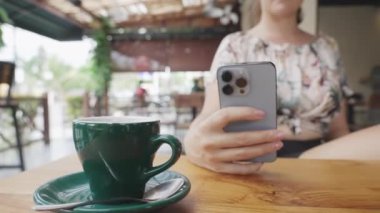 Çiçekli kazaklı bir kız, güneşli bir kafede, ahşap bir masada telefon ve yeşil bir fincan çayla oturuyor. Bir kız sıcak bir yaz gününde restoranda dinleniyor. Dolly vurdu. Yavaş çekim.