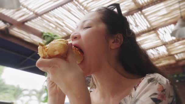 Dolly shot. Beautiful Asian girl with long hair is eating a delicious hot dog in a sunny cafe. Close-up girl is sitting in a bright sun-drenched restaurant and eating a big juicy hot dog. Slow motion — стоковое видео