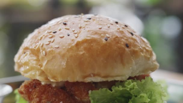 Dolly fired. Close-up of an appetizing juicy burger with crispy bun on steel tray. Golden bun with sesame fish burger, with green salad and sauce. Burger on background of wonderful bokeh. slow motion — стоковое видео