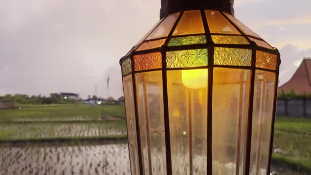 Close-up of a vintage lamp with flickering light on background of rice fields at sunset with pink sky. Antique lamp with beautiful pattern and on background of tropical landscape Bali island — 图库视频影像