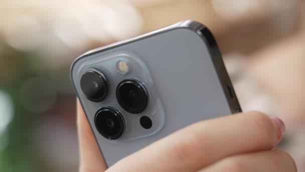 Dolly erschossen. Nahaufnahme Makroplan der Kameralinsen des neuen Apple iphone 13 Pro in der Farbe Blue Sierra vor dem Hintergrund der schönen Bokeh-Defokussierung. Moderne modische Telefon in den Händen der Person. — Stockvideo