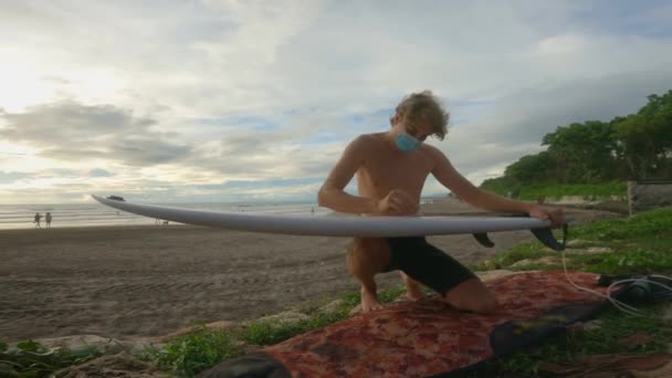 Un jeune surfeur en short et un masque médical frotte une planche de surf blanche avec de la cire sur le fond de la plage. Un surfeur masculin pendant une pandémie prépare sa fille à surfer sur les vagues. — Video