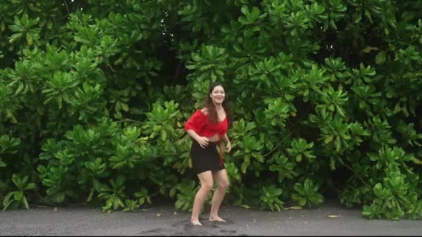 Seorang gadis Asia muda yang ceria dengan rambut panjang, blus merah dan rok mini hitam, melompat ke belakang dinding besar fajar di pantai Bali. Pelancong perempuan tersenyum dengan sukacita dan melompat dalam gerakan lambat — Stok Video