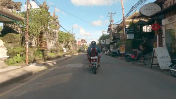 ウブド、バリ、インドネシア- 2021年12月10日:パンデミックの間、寺院、お店、バイクの間のウブド通りでバイクに乗っている人のポフ。青と晴れた日にウブドの観光センター — ストック動画