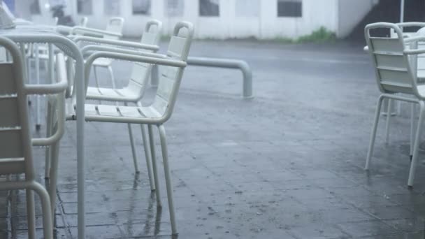 Białe stoły i krzesła nowoczesnej ulicznej restauracji w ulewnym deszczu. Puste stoły i krzesła w zamkniętej restauracji z powodu pandemii i kwarantanny koronawirusów. Chmurny dzień z deszczem. — Wideo stockowe