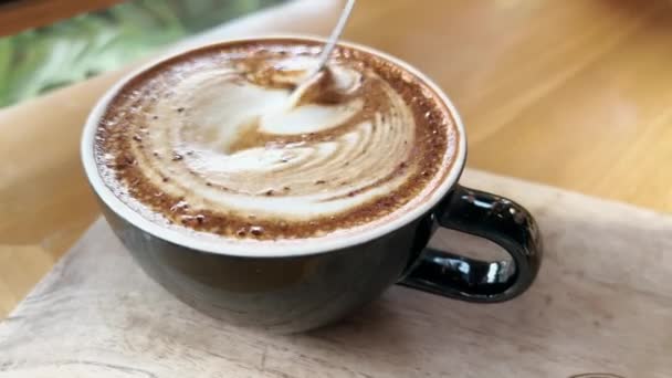 Close-up of fragrant cappuccino coffee with a beautiful latte art pattern in form of a leaf. Person is stirring with an iron spoon a delicious coffee drink standing on a wooden table in a restaurant. — Stockvideo