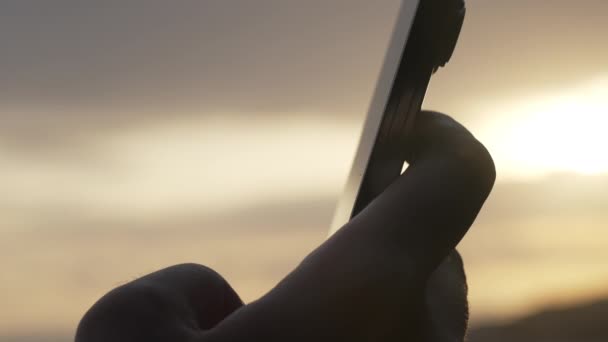 Ένα κοντινό πλάνο ενός νέου μοντέρνου τηλεφώνου στα χέρια ενός ανθρώπου στο φόντο ενός υπέροχου ηλιοβασιλέματος σε χρυσές ακτίνες φωτός. — Αρχείο Βίντεο