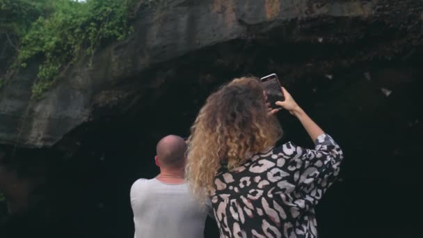 Ein Mädchen und ein Mann drehen mit ihren Handys Videos von einer riesigen Felshöhle, aus der Fledermäuse herausfliegen. Tagsüber Zeitlupe. Ein paar Reisende fotografieren fliegende Fledermäuse. — Stockvideo