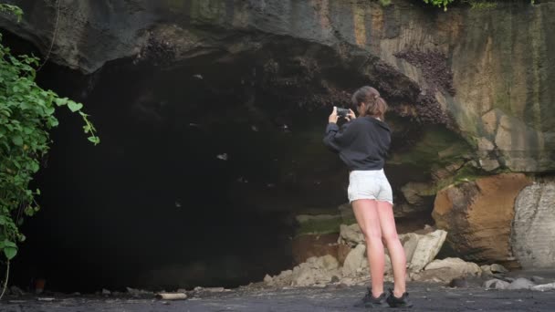 Girl stands in front of entrance to a large cave from which bats fly out and takes video on her phone. A woman in denim shorts records a video of bats on island of Bali in Indonesia. slow motion. — Vídeo de Stock