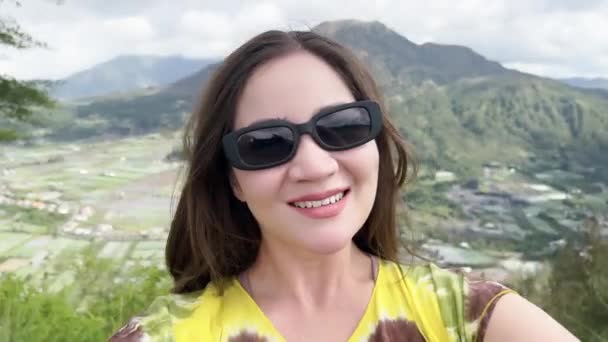 Una hermosa chica japonesa en gafas de sol de moda toma fotos de sí misma en su teléfono y sonríe en el fondo de un gran valle con un gran volcán y un cielo azul con nubes blancas. — Vídeos de Stock