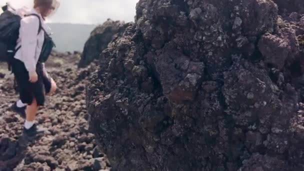 A traveler girl with sunglasses and two backpacks approaches a stone block of black lava on which a sculpture of a tower stands from small pebbles. The woman reached a new attraction. — Stockvideo