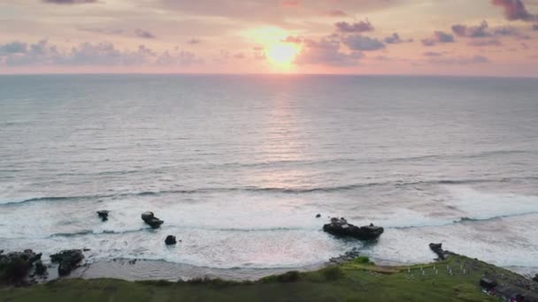 Een prachtig landschapsbeeld van een drone die over de kustlijn van donkere oceaanriffen vliegt naar een prachtige zonsondergang met een oranje zon, roze lucht en wolken. Een vogel-oog uitzicht op de zee met golven. — Stockvideo