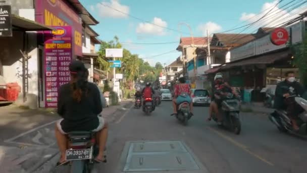 ウブド、バリ、インドネシア- 2021年12月10日:パンデミックの間、店、車、オートバイの間でウブドの通りでバイクに乗っている男のポフ。青い空と晴れた日にウブドの観光センター — ストック動画