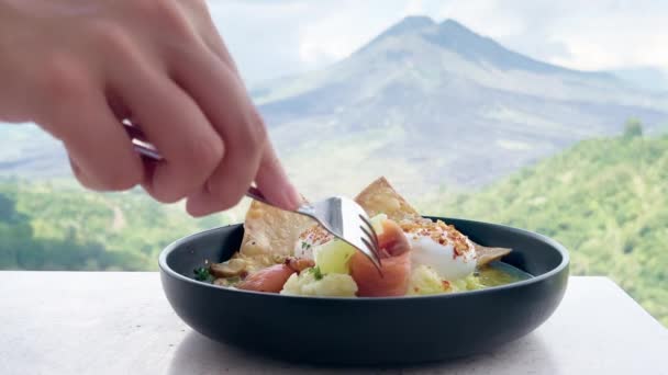 Person takes piece of salmon with fork from egg benedict morning breakfast against background of huge beautiful volcano and sky with clouds. Breakfast in a view restaurant with a view of huge — Vídeo de Stock