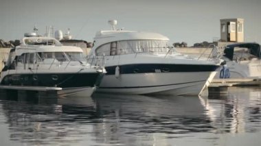 Two modern yachts, white with blue stripes, are moored at the yacht club in the beautiful golden sunset light. Beautiful boats are parked in the bay in the summer and swing on small waves.