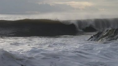 At the dark rocky shore, white sea foam is bubbling and a large wave is working and falling with great force, turning into huge white foam splashes against the blue sky. Slow motion. Bali. Indonesia.