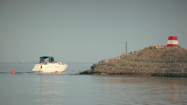 A modern white boat sails from port to sea following a route fenced with orange buoys. Boat sails behind stone ridge following the fairway. Seascape with a stone ridge boat, seabirds and a red tower. — Vídeo de Stock