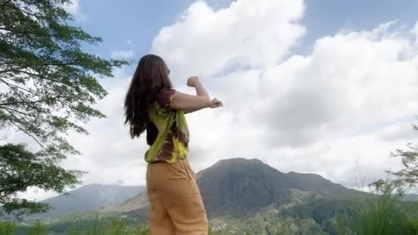 Hippie girl with long hair in colored tie dye T-shirt and yellow pants dances against background of large mountain and blue sky with clouds on sunny day. Traveler dances and enjoys nature in — Vídeo de stock