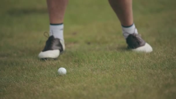 A person in white and brown golf shoes hits a white ball with a club on a green golf course. Close-up of hitting a ball with a club while playing golf on a sunny day on green grass. — 图库视频影像