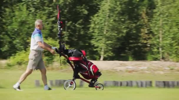 Sestroretsk, Saint-Petersburg, Russia - Jule 2 , 2012: Adult male golfer in a blue and white T-shirt and shorts walks along green golf course and rolls cart for clubs in front of him on a sunny day. — Stok video