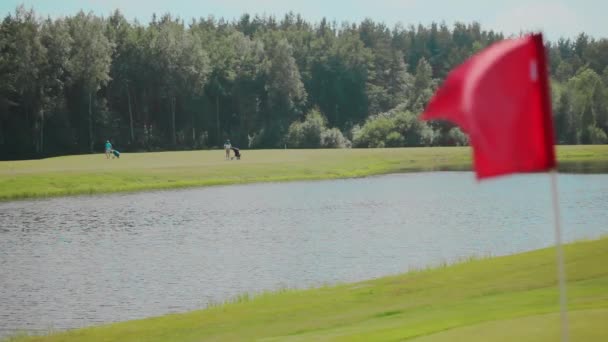 Közelkép a piros zászló integetett a szél a golfpályán tó és két golfozó hátterében zöld erdő és kék ég. Golf pálya mellett folyó egy napsütéses napon. Golfpálya tájképe — Stock videók