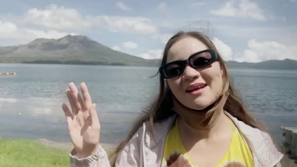 Una chica asiática en gafas de sol de moda mira a la cámara y las olas contra el fondo del lago y el gran volcán con cielo azul y nubes. Chica viajero alegre en el fondo de la montaña en el día. — Vídeos de Stock