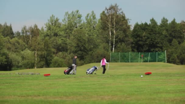 Мужчина и женщина в розовом жилете ходят по полю для гольфа и несут тележки с клюшками в солнечный день. Летний гольф в прохладную погоду. — стоковое видео