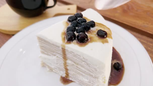 Close-up de um homem com uma colher de chá quebra um pedaço de bolo branco salgado com bagas. Uma maravilhosa sobremesa branca de neve com mirtilos e em um pires branco e uma mesa de madeira. Deliciosa torta doce. — Vídeo de Stock