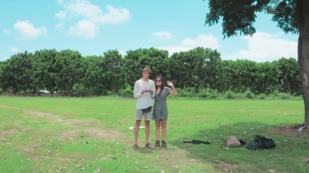 Para w okularach robi sobie selfie na dronie. Dziewczyna fale przed kamerą i facet trzyma zdalne sterowanie i kontroluje drona, odlatuje od siebie ujawniając wspaniały krajobraz z zielonymi drzewami. — Wideo stockowe
