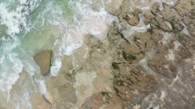 Yeşil okyanus dalgalarının beyaz köpüklü kahverengi kayalara çarptığı dramatik hava sahnesi. Güçlü enerji ve vahşi doğanın insansız hava aracı görüntülerini yükseltiyor. Hassas renkleri olan sahil manzarası.
