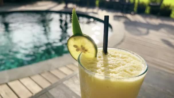 Macro close-up delicioso suco tropical com fatia de limão em vidro no belo restaurante com piscina criando brilho de luz solar. Cães de sol em ondulações de água atrás do suco de abacaxi saboroso. — Vídeo de Stock