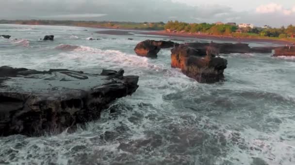 Uma bela vista de pássaros-olho de voar em torno das rochas negras no oceano na hora de ouro. Fotografias de drones da costa tropical ao pôr-do-sol. espuma do mar branco quebra em um recife escuro na ilha de Bali. — Vídeo de Stock