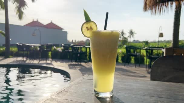 Sumo tropical delicioso com fatia de limão em vidro no belo restaurante perto de campos de arroz com piscina criando brilho de luz solar. Cães de sol em ondulações de água atrás do suco de abacaxi saboroso. — Vídeo de Stock