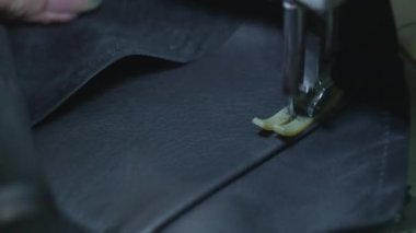 Dikiş makinesindeki siyah deri ceketin iğnesini dikmeye yakın plan. Terzi kadın deri eşyalar yapıyor, karanlık bir fabrika odasında bir sıra dikiyor..