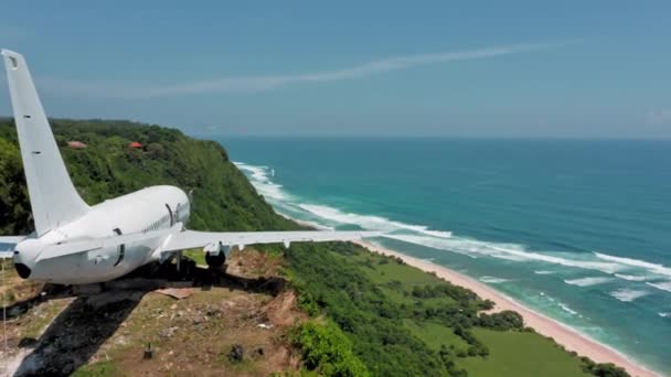 Bílé letadlo na okraji útesu v zeleném tropickém ráji u oceánu. Opuštěné letadlo na ostrově Bali s panoramatickým výhledem na oceán na pláži. Letecký pohled na letadlo a kopce nad mořským pobřežím. — Stock video