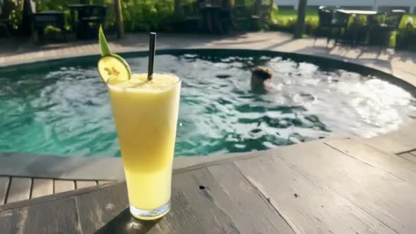 Close-up delicioso suco tropical com fatia de limão em vidro no belo restaurante terraço com piscina criando brilho de luz solar. Sumo de abacaxi saboroso atrás do qual uma menina está nadando na piscina — Vídeo de Stock