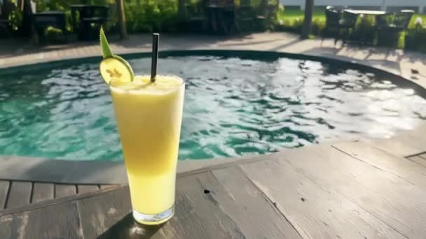 Close-up delicioso suco tropical com fatia de limão em vidro no belo restaurante terraço com piscina criando brilho de luz solar. Cães de sol em ondulações de água atrás do suco de abacaxi saboroso. — Vídeo de Stock