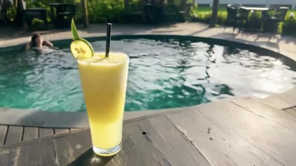 Close-up delicioso suco tropical com fatia de limão em vidro no belo restaurante terraço com piscina criando brilho de luz solar. Sumo de abacaxi saboroso atrás do qual uma menina relaxa na piscina. — Vídeo de Stock