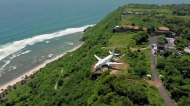 Okyanus yakınlarındaki yeşil tropikal cennette uçurumun kenarındaki uçağa zum yap. Plajda panoramik okyanus manzaralı Bali adasında terk edilmiş bir uçak. Deniz kıyısındaki uçak ve tepelerin havadan görüntüsü. 4k