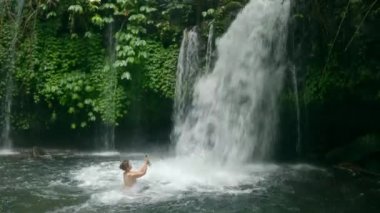 Çılgın bir kadın blogcu, tropikal nehirde büyük bir şelalenin altında telefonuyla video çekiyor. Modern cep telefonu kamerasıyla su altında beyaz tenli bir kız. Bali 'de eğlenen kadın