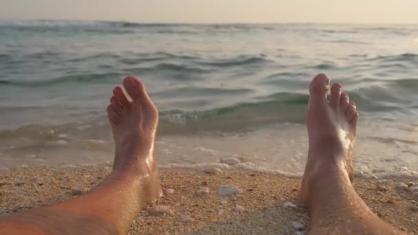 Zbliżenie pięknych męskich nóg na tropikalnym wybrzeżu z małymi falami oceanicznymi o zachodzie słońca. Mężczyzna lubi surfować na plaży, a fale morskie uderzają mu w stopy latem. Zwolniony ruch. — Wideo stockowe