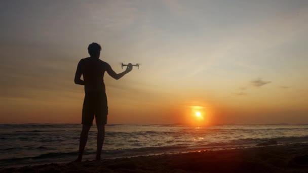 La silueta de un hombre en pantalones cortos sobre el fondo del océano y el cielo del atardecer lanza un dron de su mano hacia un paisaje maravilloso con el sol y un cielo del tabaco — Vídeo de stock