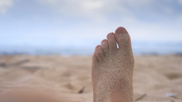 Een man beweegt zijn tenen tijdens het ontspannen op een strand met zand en blauwe lucht tijdens de dag. Close-up van menselijke tenen besprenkeld met zand. Pov van een persoon genieten van een vakantie op het strand en kijken naar zijn been. — Stockvideo