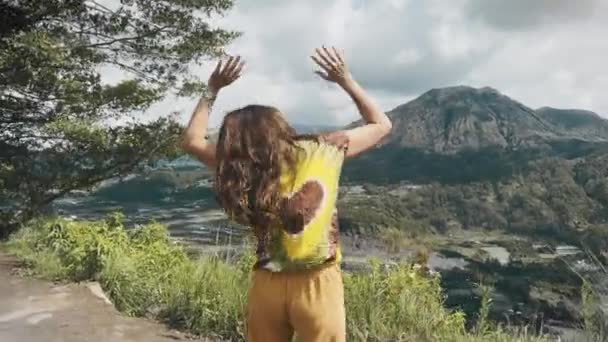Chica hippie con el pelo largo y oscuro en colorido corbata camiseta y pantalones amarillos baila contra el fondo de la gran montaña y el cielo azul con nubes en el día. Viajero baila y disfruta de la naturaleza en las montañas — Vídeo de stock