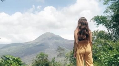 Sarı pantolonlu, uzun saçlı esmer kız dağın tepesinde duruyor ve beyaz bulutlu dev volkana ve gökyüzüne bakıyor ve ellerini kaldırıyor. Kız sabahları esniyor. 4k.