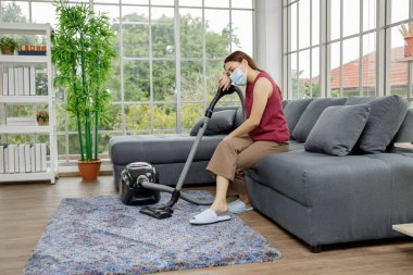 Asyalı ev hanımı kanepede ve elektrikli süpürgede oturuyor. 
