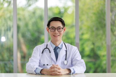 Beyaz üniformalı, göz çimeni takan Asyalı doktor portresi oturuyor ve arka planda gülümsüyor. 