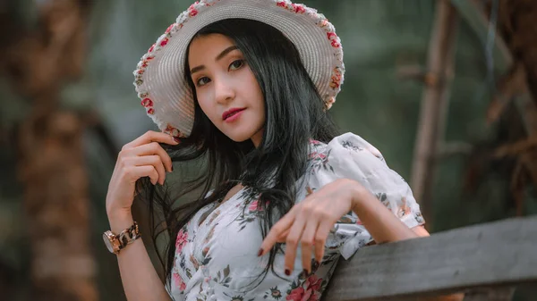 美丽的年轻亚洲女人 穿着复古的花朵 粉色服装风格和帽子复古风格 坐在公园背景的木桥上 看着相机 度假时的自然光肖像和复古风格的理念 — 图库照片