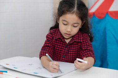 Ekose gömlekli bebek Asyalı kız odadaki beyaz masaya oturup kağıt kitap çiziyor.